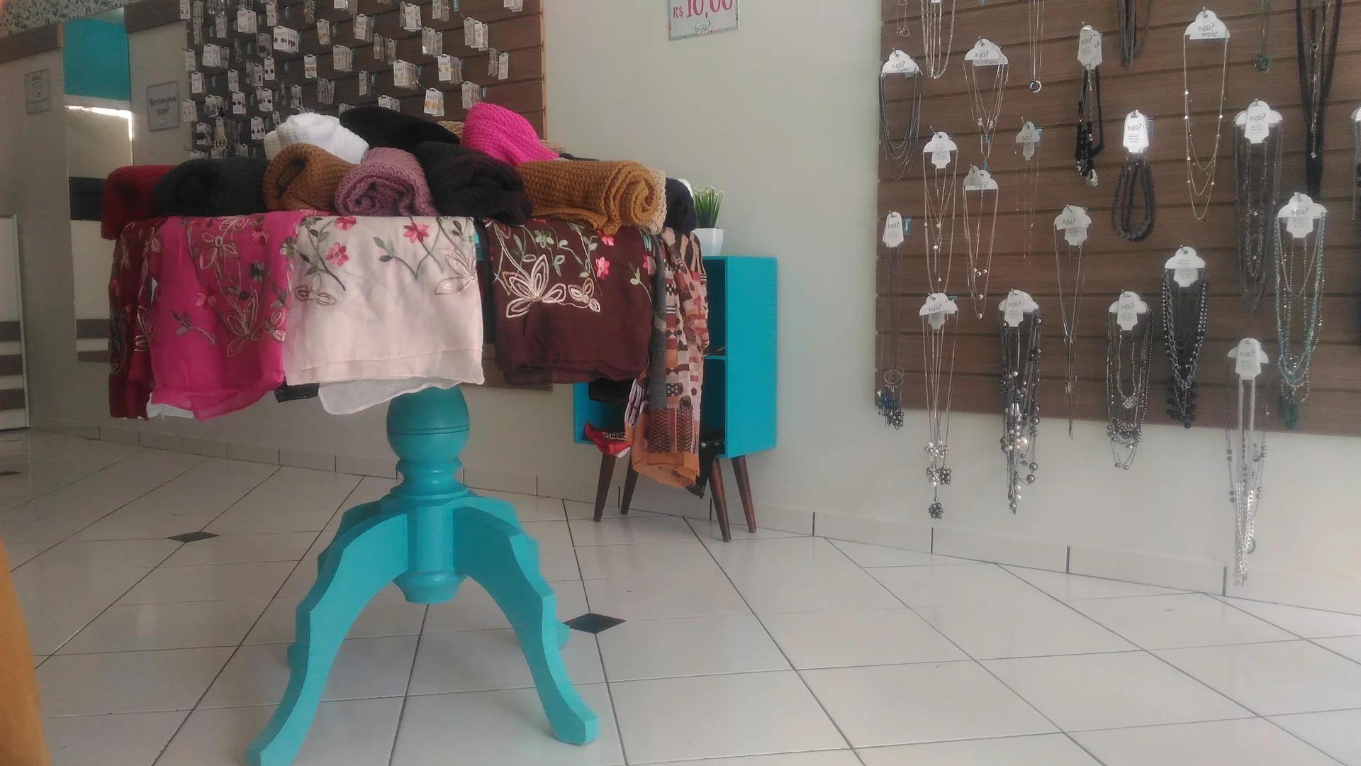 Interior da Ouvi Biju com mesa turquesa expondo lenços e painel canaletado com colares, Limeira — DOA Estúdio